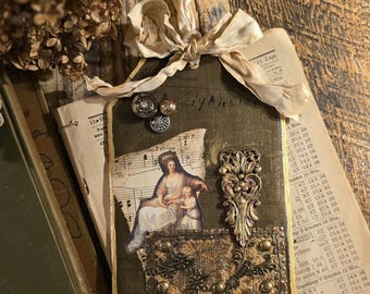May include: A vintage-style tag with a collage of elements. It features a printed image of a woman and child, sheet music, and ornate metal embellishments. The tag is adorned with buttons and a cream-colored ribbon.