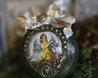 May include: A green, round decorative ornament with a gold-framed image of an angel in a yellow robe. The ornament has a gold button and cream-colored ribbon at the top. The ornament is sitting on top of a book.