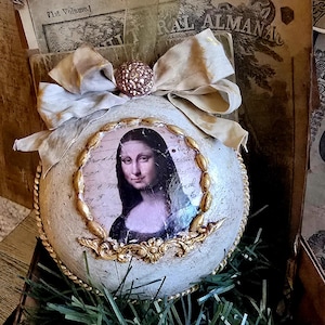 Op de afbeelding: Ornament met een afdruk van de Mona Lisa, omlijst met gouden filigraan, tegen een getextureerde, gebroken witte ondergrond. Het ornament is versierd met een crèmekleurige strik en een decoratieve gouden bal. Vintage boeken en groen maken het geheel compleet.