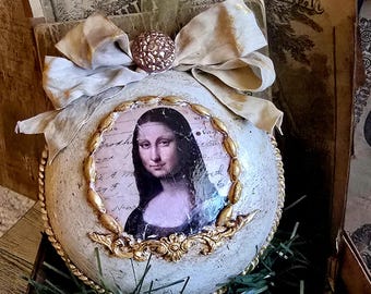 May include: Ornament featuring a print of the Mona Lisa, framed with gold filigree, set against a textured, off-white surface. The ornament is adorned with a cream-colored bow and a decorative gold ball. Vintage books and greenery complete the display.