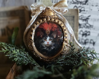 May include: Ornate, gold-colored oval ornament featuring a portrait of a cat with black and white fur, a red headdress, and a gold frame. The ornament is adorned with a cream-colored bow and sits atop greenery, evoking a vintage aesthetic.