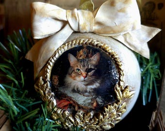 May include: A decorative Christmas ornament featuring a regal cat portrait. The round ornament has a gold-colored frame and a cream-colored background, topped with a large, cream-colored bow. The cat wears a crown and is surrounded by gold leaf accents.