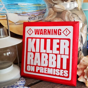 Könnte beinhalten: Rotes Holzschild mit weißem Text, der "WARNUNG! TÖDLICHER HASE AUF DEM GELÄNDE" lautet. Das Schild hat auf beiden Seiten des Wortes "WARNUNG!" einen weißen Ausrufezeichen in einer Raute.