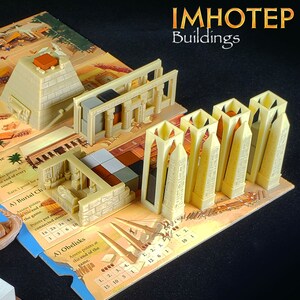 May include: A close-up of a board game called "Imhotep: Buildings". The game features a variety of buildings, including pyramids, temples, and obelisks. The buildings are made of beige plastic and are arranged on a colorful game board.