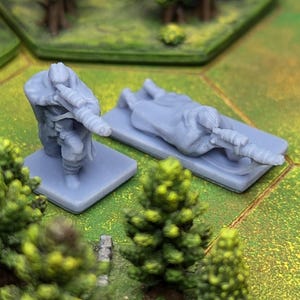 May include: Two gray miniature figures, one standing and one lying down, are positioned on small square bases. The figures appear to be soldiers holding rifles. The background features a green and brown hexagonal game board with miniature trees.