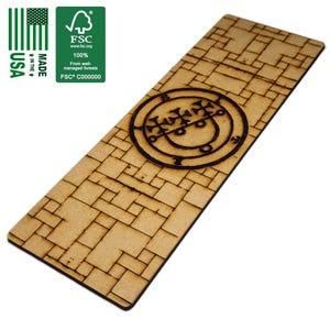 Könnte beinhalten: Rechteckiger Holz-Lesezeichen mit Backsteinmuster und schwarzem, kreisförmigem Symbol. Das Lesezeichen wird in den USA hergestellt und ist FSC-zertifiziert. Das Symbol ist ein detailliertes, dunkles Design. Das Lesezeichen ist hellbraun.