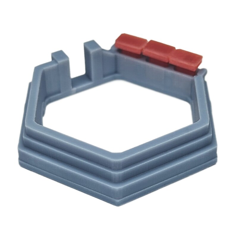 May include: A gray hexagonal-shaped plastic base with a raised lip and three red plastic pieces on top. The base is designed to hold a small object, such as a ring or a coin.