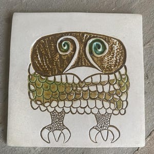 Può includere: Una piastrella quadrata in ceramica con un'illustrazione stilizzata di un gufo. Il gufo ha un piumaggio marrone testurizzato, occhi a spirale verdi e un becco bianco. Il corpo è coperto da un motivo a scaglie con smalto verde e marrone.