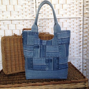 May include: A tote bag constructed from various shades of blue denim fabric pieces arranged in a patchwork pattern. The bag features two matching denim handles attached to the top, with a solid light blue denim base and a rectangular body shape.