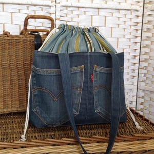 May include: A tote bag constructed from dark blue denim fabric featuring two exterior back pockets with orange stitching and a small red tag. The bag includes a drawstring closure made of striped green, yellow, and white fabric with white cords.