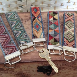 May include: Five fabric key fobs with denim backing and woven geometric patterns in red, green, blue, and orange. Each strap features a silver-toned metal clamp and a circular split ring. One key fob is attached to a single brass-coloured metal key.
