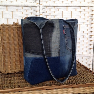 May include: A patchwork denim tote bag with a dark blue denim handle and a red Levi's tag. The bag is made from various shades of blue denim. A small wicker basket is to the left of the bag.