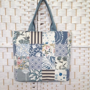 May include: A patchwork tote bag with a denim handle. The bag features a variety of blue and white floral and geometric patterns. The bag is hanging against a white woven background.