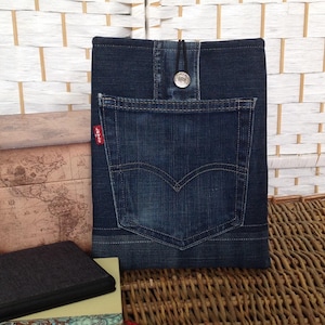 May include: A dark blue denim pouch featuring a single back pocket with a signature Levi's red tab on the side. The pouch is secured with a silver-toned metal button and a black elastic loop closure centered at the top edge of the fabric.