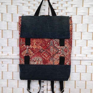 May include: A rectangular backpack featuring a red and orange patterned fabric centre panel with a dark blue denim top flap and bottom base. The bag includes black fabric straps, silver metal hardware clips, and adjustable black shoulder straps.