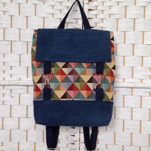 May include: A backpack featuring a geometric triangle pattern in shades of red, orange, yellow, green, and blue. The bag has a dark blue denim-textured flap and base, dark blue fabric straps, silver-toned metal hardware, and a top handle.