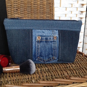 May include: A denim makeup bag with a pocket detail. The bag is made from various shades of blue denim and features a zip closure. A makeup brush and other cosmetic items are visible in the foreground.