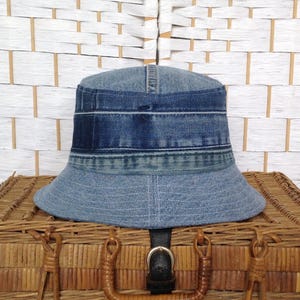 May include: A bucket hat constructed from repurposed blue denim fabric. The hat features a patchwork design with varying shades of blue, including a darker band around the crown and a lighter denim brim, showcasing visible seams and stitching details.