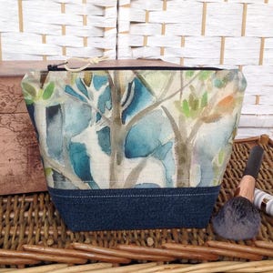 May include: A rectangular cosmetic bag with a watercolor forest scene featuring a white deer. The bag has a dark blue denim base and a zipper closure. Makeup brushes and a small container are next to the bag.