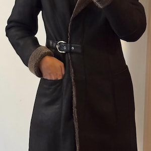 May include: A black shearling coat with a brown fur collar and cuffs. The coat features a belt at the waist and a classic design. The coat has a long length and a tailored fit, suitable for cold weather.