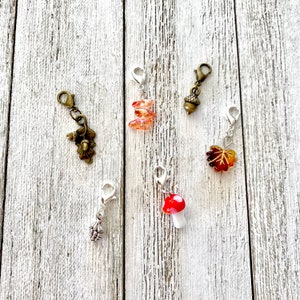 May include: Five different charm clips with autumnal themes. The charms include a mushroom, an acorn, a leaf, and two stacked glass beads. The charms are attached to silver-toned clips.