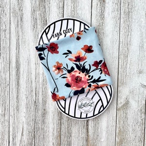 May include: A pale blue fabric with a floral design of pink and rust-coloured flowers and black stems. The fabric is folded and placed on a white and black striped oval object. The text "YARN COZY" is visible.