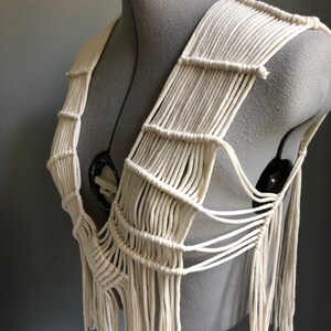 May include: Cream-colored macrame vest displayed on a mannequin. The vest features a woven design with vertical and horizontal rope elements, creating a fringed bottom. The neutral color and intricate knotting suggest a bohemian or handcrafted style.