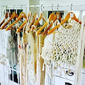 May include: A collection of cream-colored macrame and crochet garments hanging on wooden hangers. The garments include tops, dresses, and vests with intricate woven patterns and fringe details. The display is set against a white backdrop.