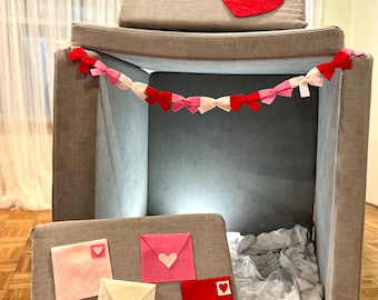 May include: A gray fabric playhouse with a red heart on the top and a pink and red heart garland inside. The playhouse has a white fabric floor and four pink and red felt envelopes with hearts on them.
