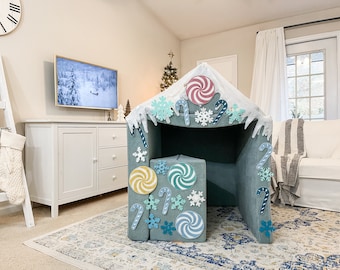 May include: A blue and white playhouse tent decorated with candy canes, snowflakes, and peppermint swirls. The tent is set up in a living room with a Christmas tree in the background.