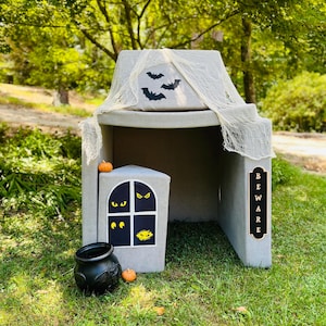 Può includere: Una casetta di gioco grigia a forma di casa infestata con un calderone nero, zucche arancioni e un cartello che dice "BEWARE".