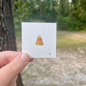 May include: A small watercolor painting of a candy corn on a square piece of white paper. The candy corn is painted with orange, yellow, and white colors. The painting is signed with the initials "TZ".