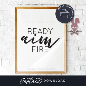 May include: A framed printable with the text "READY aim FIRE" in black script font on a white background. The frame is brown wood. The image is on a white brick wall with a pink skull and crossbones graphic in the upper right corner.