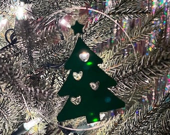 May include: A green Christmas tree ornament with a star on top and three hearts cut out of the design. The ornament is hanging on a Christmas tree with lights.