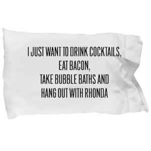 May include: White pillowcase with black text that reads "I just want to drink cocktails, eat bacon, take bubble baths and hang out with Rhonda."