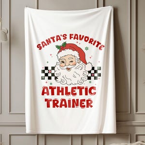 Könnte beinhalten: Weiße Kuscheldecke mit einer roten und schwarzen Grafik von Weihnachtsmann mit dem Text "Santas Lieblings-Fitness-Trainer".