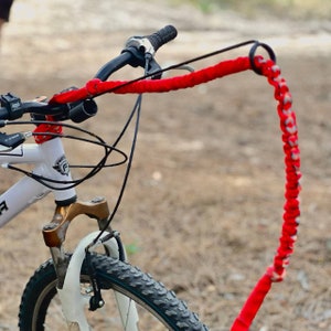 May include: A white bicycle with a black handlebar and a red braided paracord leash attached to the center. The leash features a circular metal ring at the end and a woven pattern. The bicycle has a front suspension fork and a knobby black tire.