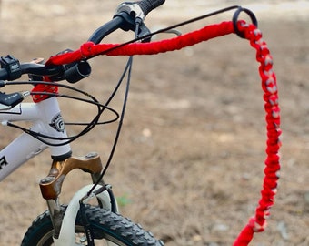 May include: A white bicycle with a black handlebar and a red braided paracord leash attached to the center. The leash features a circular metal ring at the end and a woven pattern. The bicycle has a front suspension fork and a knobby black tire.
