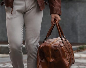May include: A brown leather jacket with quilted shoulder detailing, a gray mock neck shirt, light beige trousers, and a brown leather duffel bag. The jacket features a stand collar, zippered chest pockets, and long sleeves with zippered cuffs.