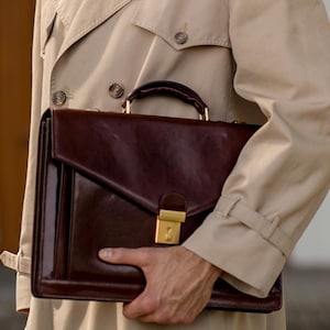 May include: A beige trench coat with a classic collar, shoulder epaulets, and button details, paired with a dark brown leather briefcase featuring a structured rectangular shape, a top handle, and a gold-toned metal clasp closure on the front flap.
