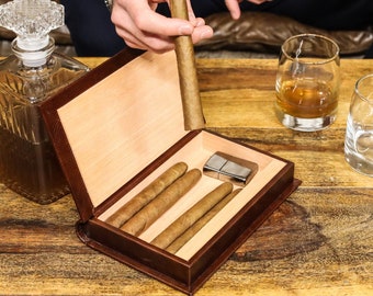 Op de afbeelding: Een houten sigarenkist met een bruine leren buitenkant en een licht houten interieur. De kist is open en bevat verschillende sigaren en een zilveren aansteker. Er staan twee glazen op de tafel, een met whisky en een leeg.