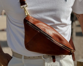 May include: A brown leather fanny pack with a gold zipper and a gold clasp. The fanny pack is worn over a white t-shirt with a small embroidered logo.