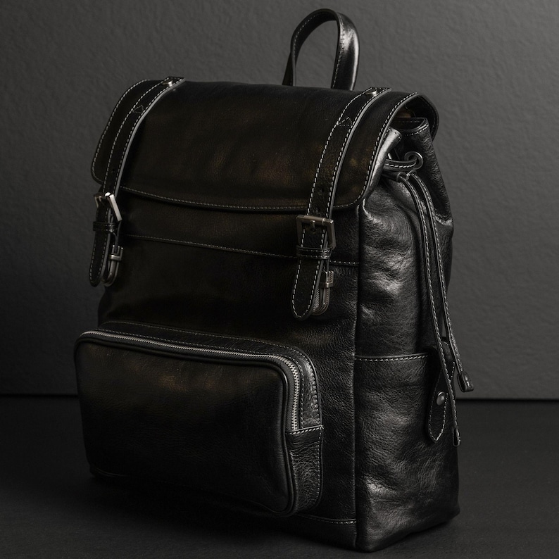 K&ouml;nnte beinhalten: Ein schwarzer Lederrucksack mit einem &Uuml;berschlag, zwei Schnallenriemen, einer vorderen Rei&szlig;verschlusstasche, einem Tragegriff und seitlichen Kordelz&uuml;gen. Der Rucksack ist mit sichtbaren wei&szlig;en N&auml;hten an den Kanten und silberfarbenen Metallbeschl&auml;gen versehen.