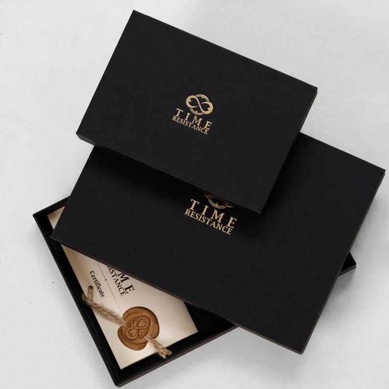 K&ouml;nnte beinhalten: Zwei gestapelte rechteckige schwarze Geschenkboxen, die jeweils das goldfarbene Logo und den Schriftzug TIME RESISTANCE tragen. Die untere Box ist leicht ge&ouml;ffnet und zeigt ein cremefarbenes Zertifikat mit einem braunen Wachssiegel.