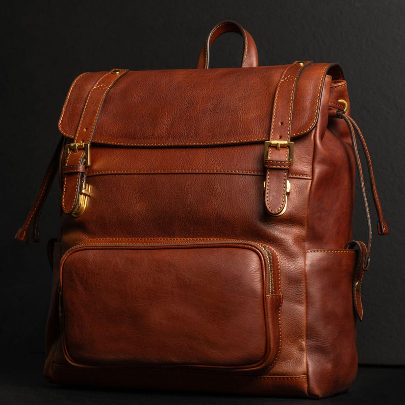 K&ouml;nnte beinhalten: Ein cognacbrauner Lederrucksack mit &Uuml;berschlagklappe, zwei verstellbaren Schnallenriemen, goldfarbenen Metallbeschl&auml;gen, einer gro&szlig;en Fronttasche mit Rei&szlig;verschluss, Seitentaschen und einem Tragegriff mit glatter, polierter Oberfl&auml;che.