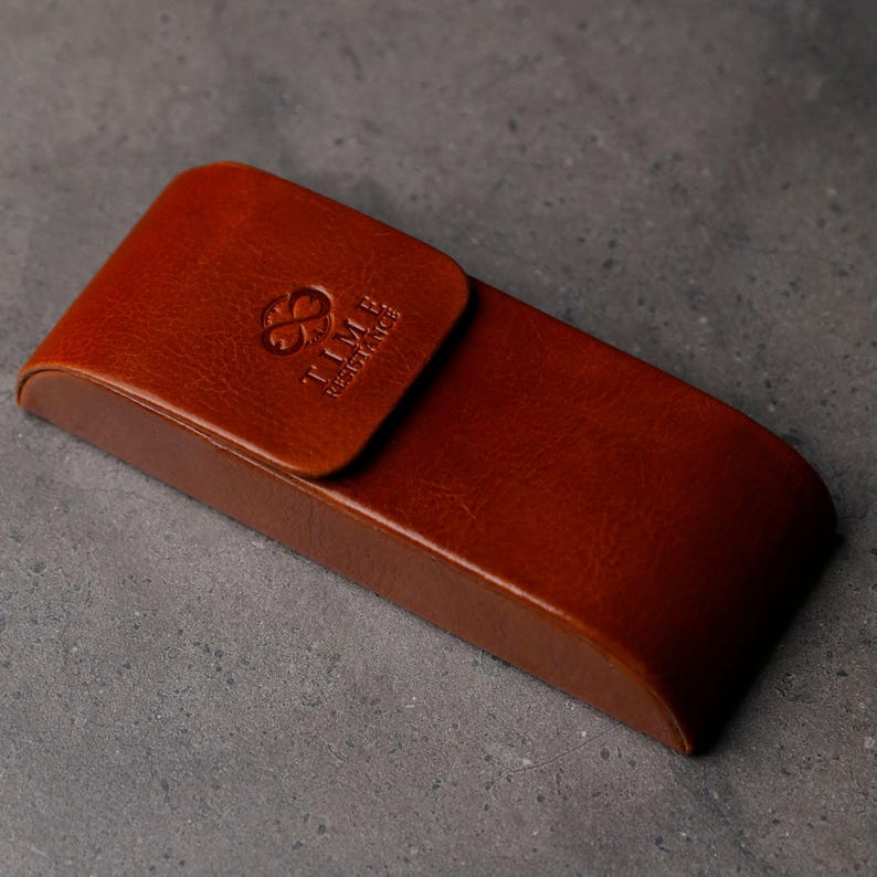 May include: A rectangular brown leather glasses case with a curved flap closure. The front of the flap features an embossed logo with the text TIME RESISTANCE. The leather has a smooth, polished finish with visible natural grain textures.