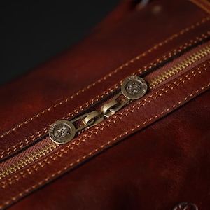K&ouml;nnte beinhalten: Nahaufnahme einer braunen Ledertasche mit einem goldenen Rei&szlig;verschluss und zwei Metall-Zippern. Die Zipper haben ein Logo mit einer stilisierten Fleur-de-lis.