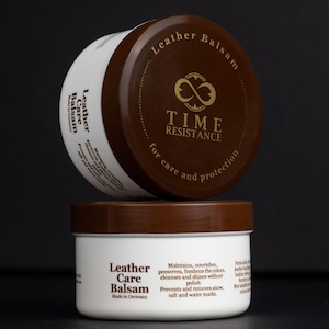 May include: A white cylindrical container of Time Resistance Leather Balsam with a brown lid. The lid features a gold infinity logo and text. The side label includes the product name, Made in Germany, and instructions for leather care and protection.