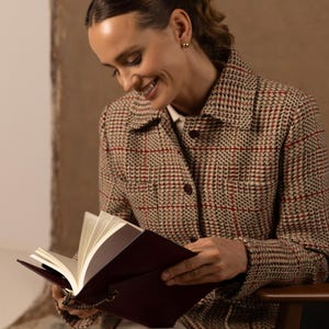 May include: A long-sleeved jacket featuring a tan, black, and red plaid pattern with a collared neckline and dark buttons. The jacket is worn over a white shirt. A dark burgundy notebook with white pages and a small metal chain is held in view.