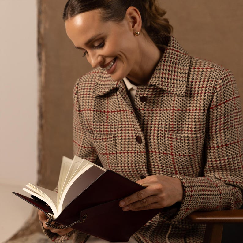May include: A long-sleeved jacket featuring a tan, black, and red plaid pattern with a collared neckline and dark buttons. The jacket is worn over a white shirt. A dark burgundy notebook with white pages and a small metal chain is held in view.